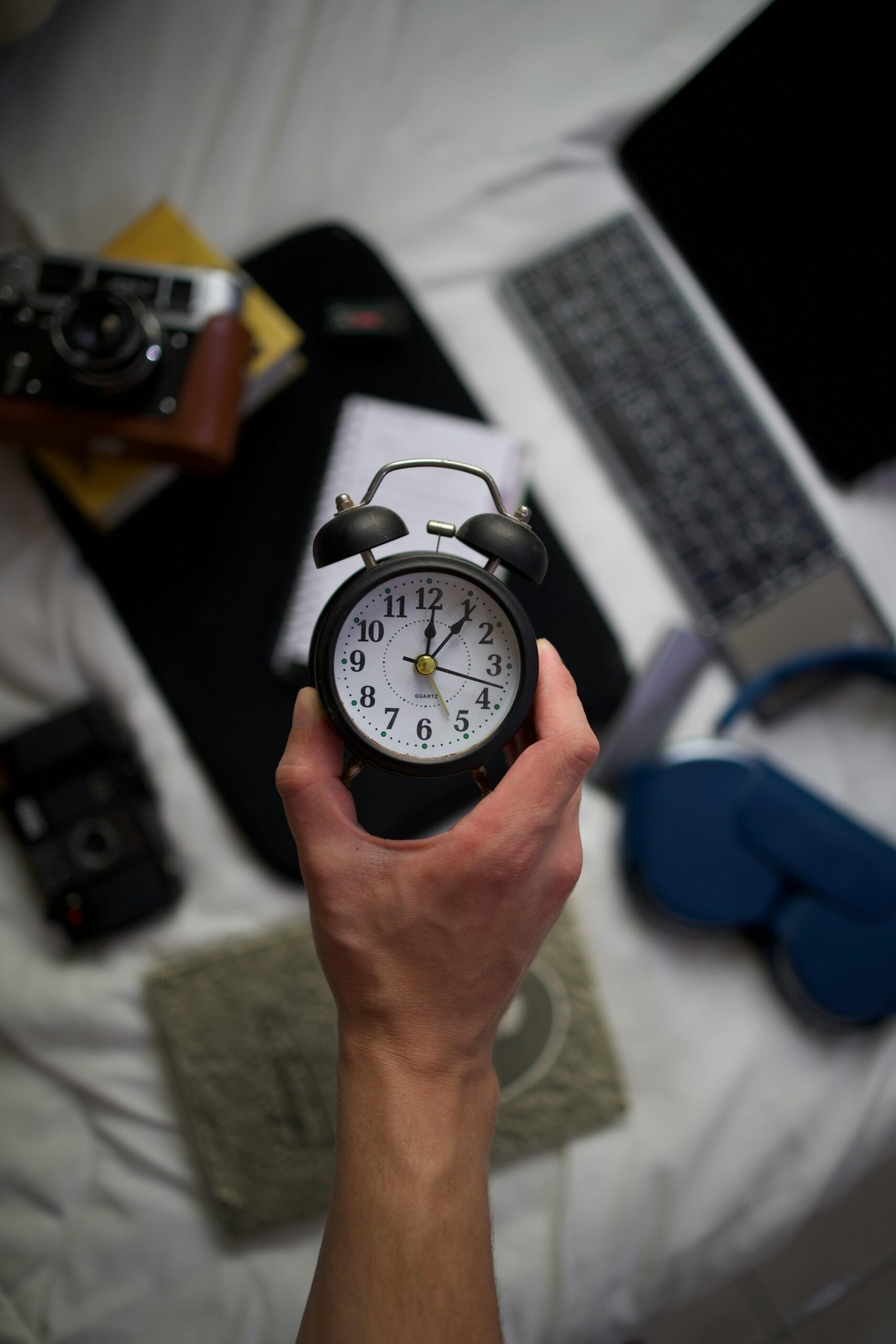 A hand holds an alarm clock over a desk with a laptop, camera, and books.