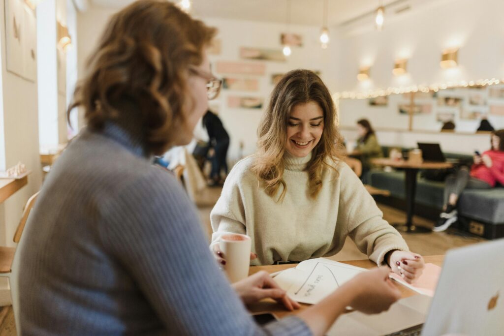 Two women in a cozy cafe discussing papers with smiles, capturing a warm and stylish ambiance.